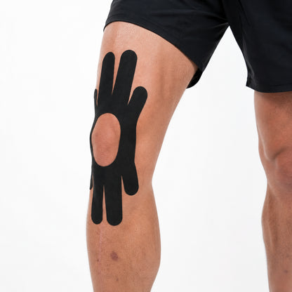 Svart förklippt kinesiologitejp applicerad runt knäskålen, visad från sidan för att tydliggöra tejpens placering.